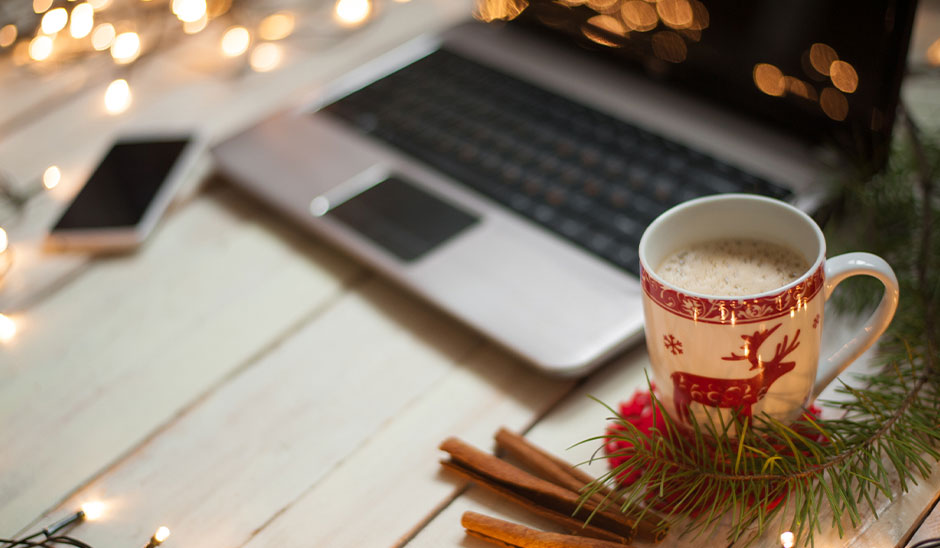 A cozy holiday workspace featuring a laptop, a mug of coffee, and cinnamon sticks on a wooden table with festive lights.