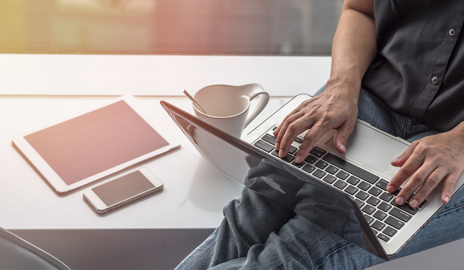Person typing on a laptop with a tablet and smartphone nearby on a table.