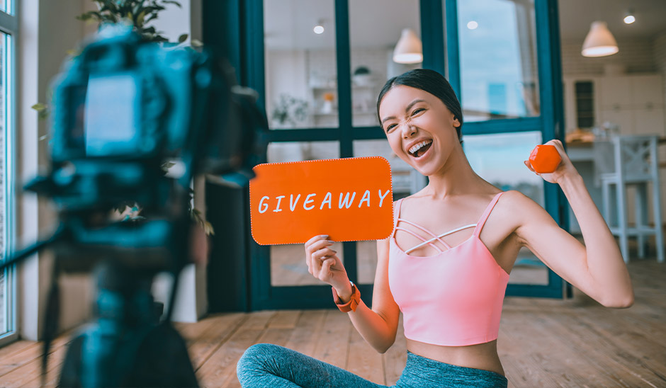 Woman with dark hair in a pink top showing a sign that says 'GIVEAWAY' while holding a dumbbell.