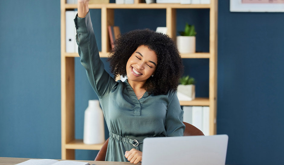Smiling woman celebrating success at a laptop in a modern office.