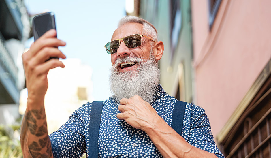Smiling elderly man with a beard taking a selfie outdoors in a vibrant shirt and sunglasses.