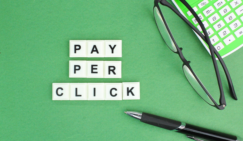 Playful arrangement of 'PAY PER CLICK' letters with glasses and a calculator on green background.