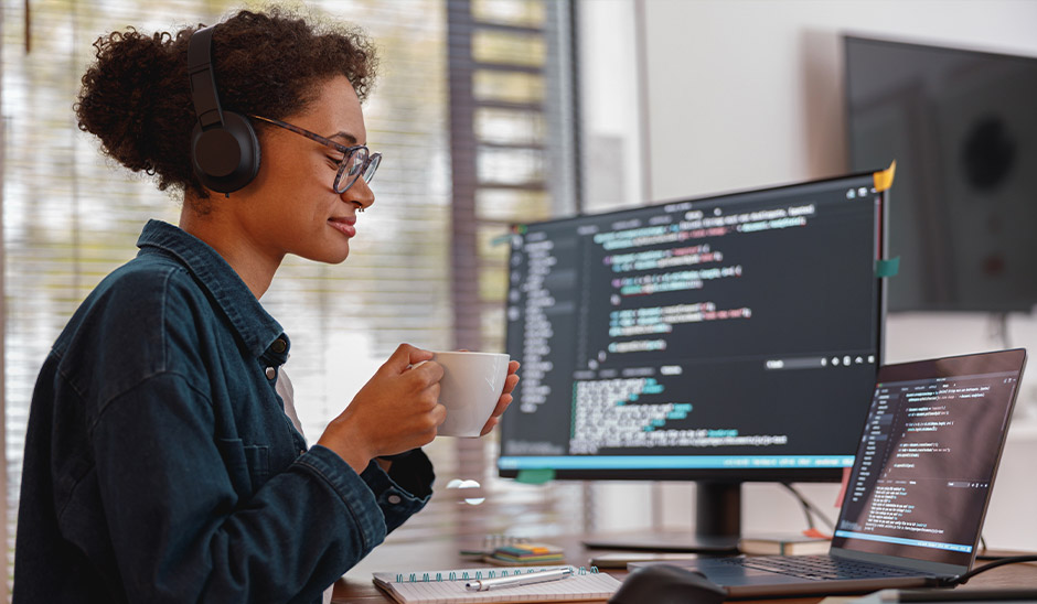 A woman with curly hair wearing headphones enjoys coffee while programming on dual monitors.