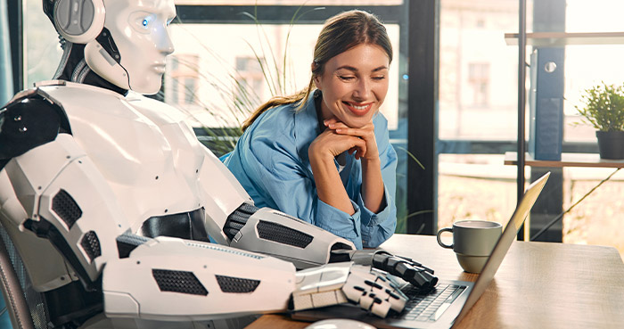 A woman smiles while watching a robot type on a laptop at a table.
