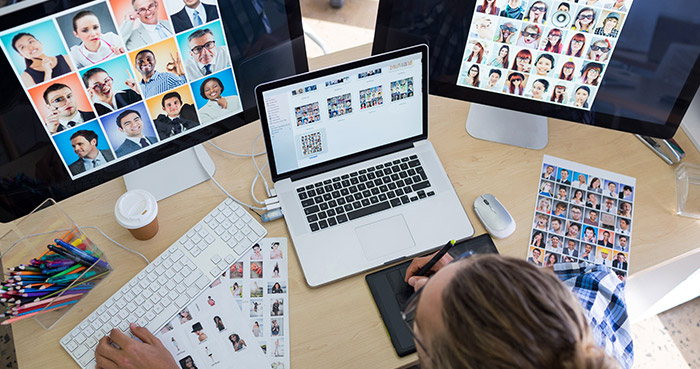 Creative workspace with a laptop and dual monitors displaying diverse portrait photos, and a person working with a graphics tablet.