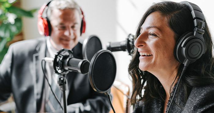 A woman with long dark hair wearing headphones smiles while podcasting with a man in a suit behind a microphone.