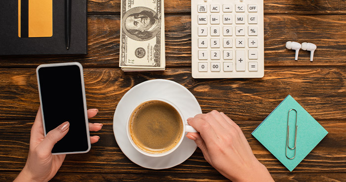 set-your-budget | Idea Stream Marketing A person holding a smartphone and a cup of coffee on a wooden table with cash, a calculator, and stationery.