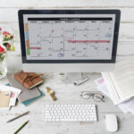 A desk setup featuring a calendar on a monitor, flowers, notebooks, and stationery.