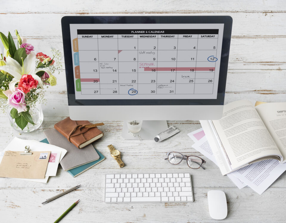 A desk setup featuring a calendar on a monitor, flowers, notebooks, and stationery.