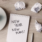 Notebook with the words 'New Year New Plans!' beside a cup of coffee and crumpled paper.