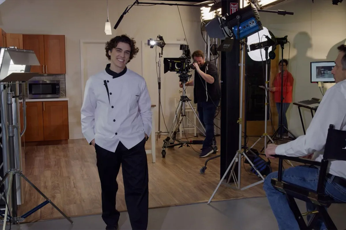 Kitchen-Area | Idea Stream Marketing A young chef in a kitchen studio smiles during a filming setup, with crew members and equipment in the background.
