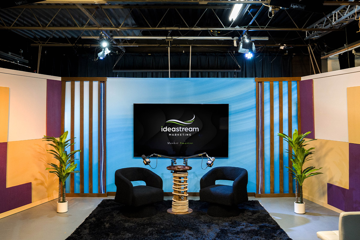 podcast-set | Idea Stream Marketing A modern podcast studio setup featuring two black chairs, a round table, a television screen with the Ideastream Marketing logo, and potted plants.