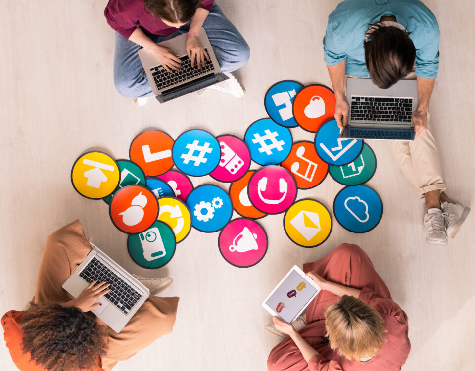 Group of people using laptops surrounded by colorful social media icons on the floor.
