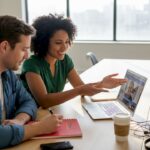 A woman explains something to a man while they watch a video on a laptop in a bright office.