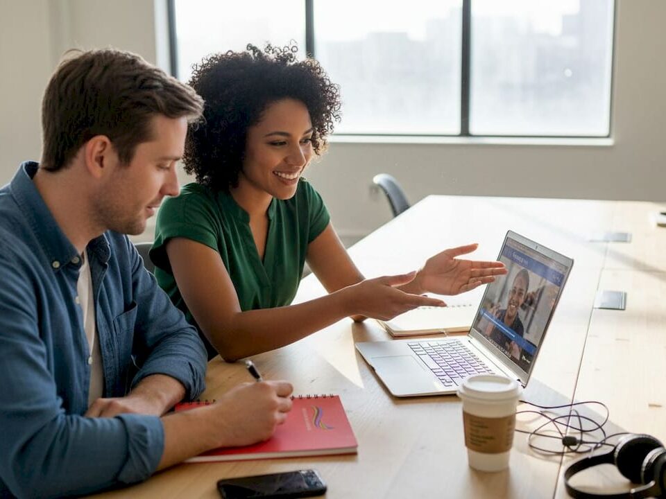 A woman explains something to a man while they watch a video on a laptop in a bright office.