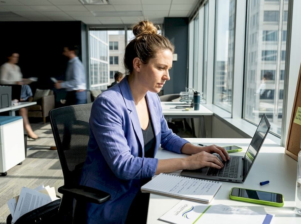 A woman types on a laptop at a sunlit office desk, with notes and phones nearby.