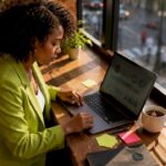 Marketer Analyzing Data in Home Office | Idea Stream Marketing A focused marketer in a bright home office analyzing data on a laptop, with sticky notes and a cup of coffee nearby.