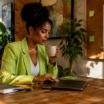 Professional woman in a bright green blazer enjoying coffee while using a laptop in a modern workspace.
