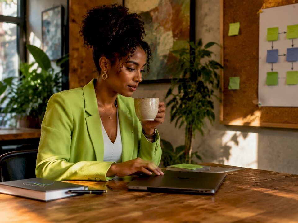 Professional woman in a bright green blazer enjoying coffee while using a laptop in a modern workspace.