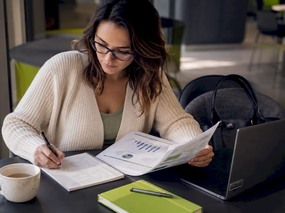 A woman analyzing a report and taking notes at a desk with a laptop and coffee.
