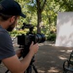 Videographer setting up a video camera on a tripod in a park with a blank backdrop.
