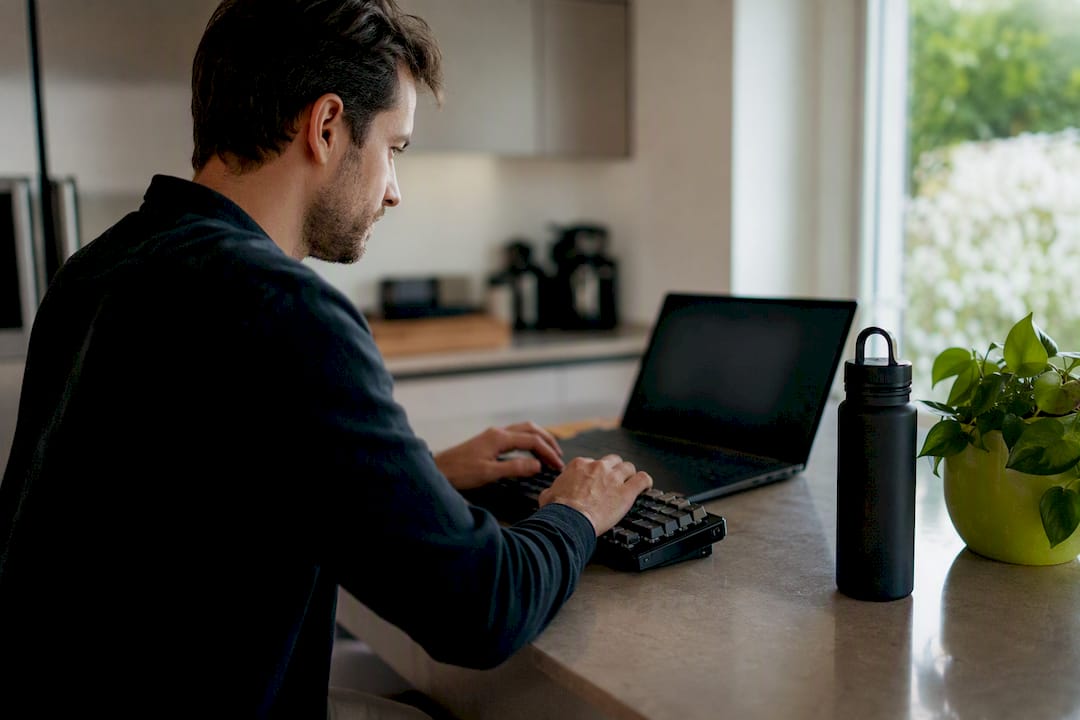 A developer typing on a keyboard at a modern kitchen table with a laptop and a plant nearby.