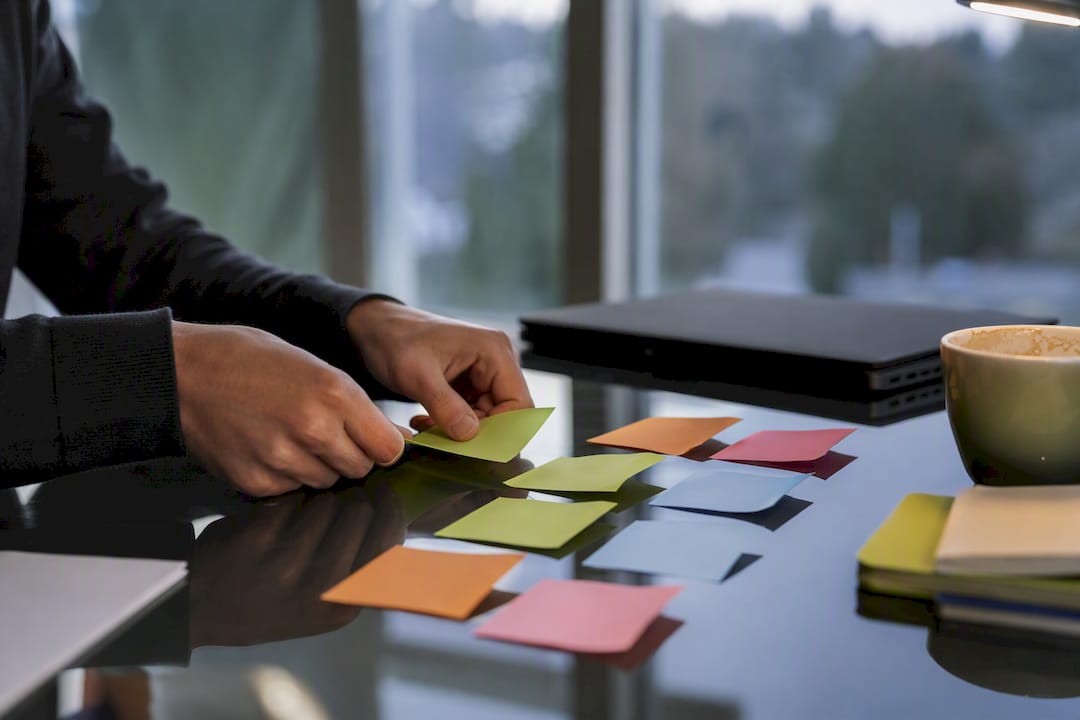 A person organizing colorful sticky notes on a glass desk, with a laptop and a coffee cup nearby.