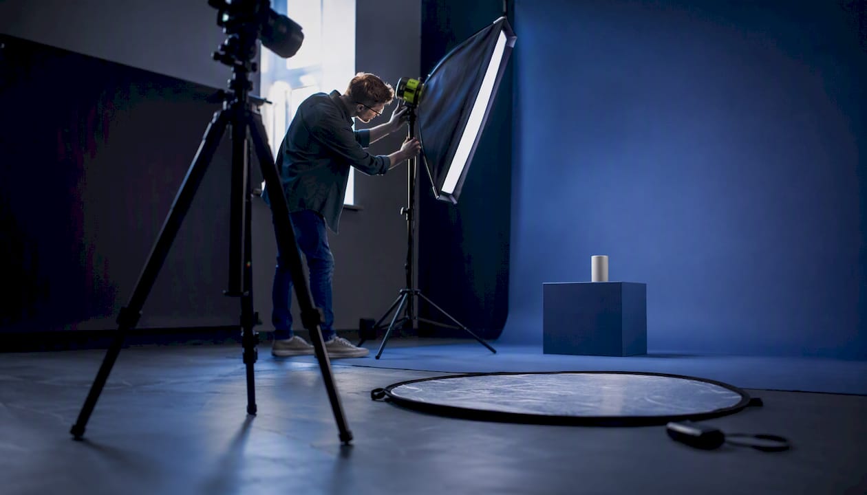 Photographer adjusting lighting in a studio with a blue backdrop and a product on a pedestal.