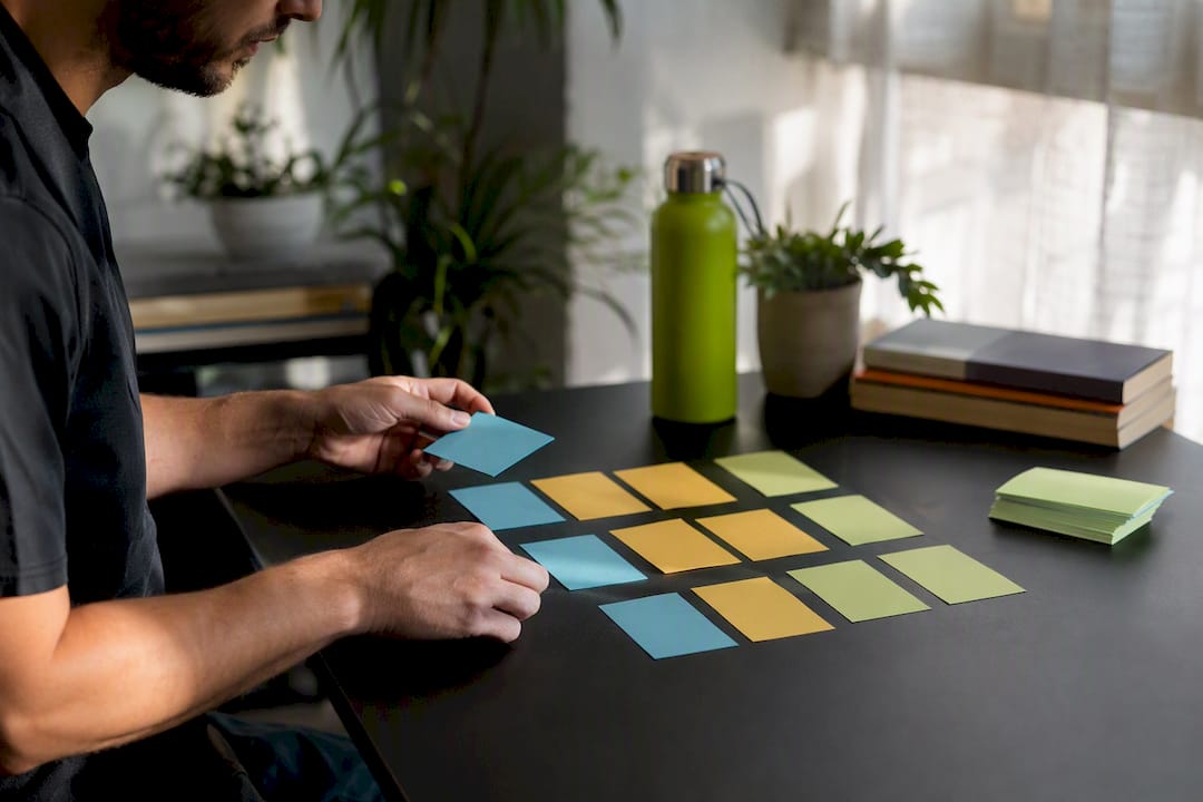 Person arranging colorful sticky notes on a black table with a water bottle and plants in the background