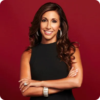 A confident woman with long dark hair wearing a black dress and earrings, posing with arms crossed against a red background.